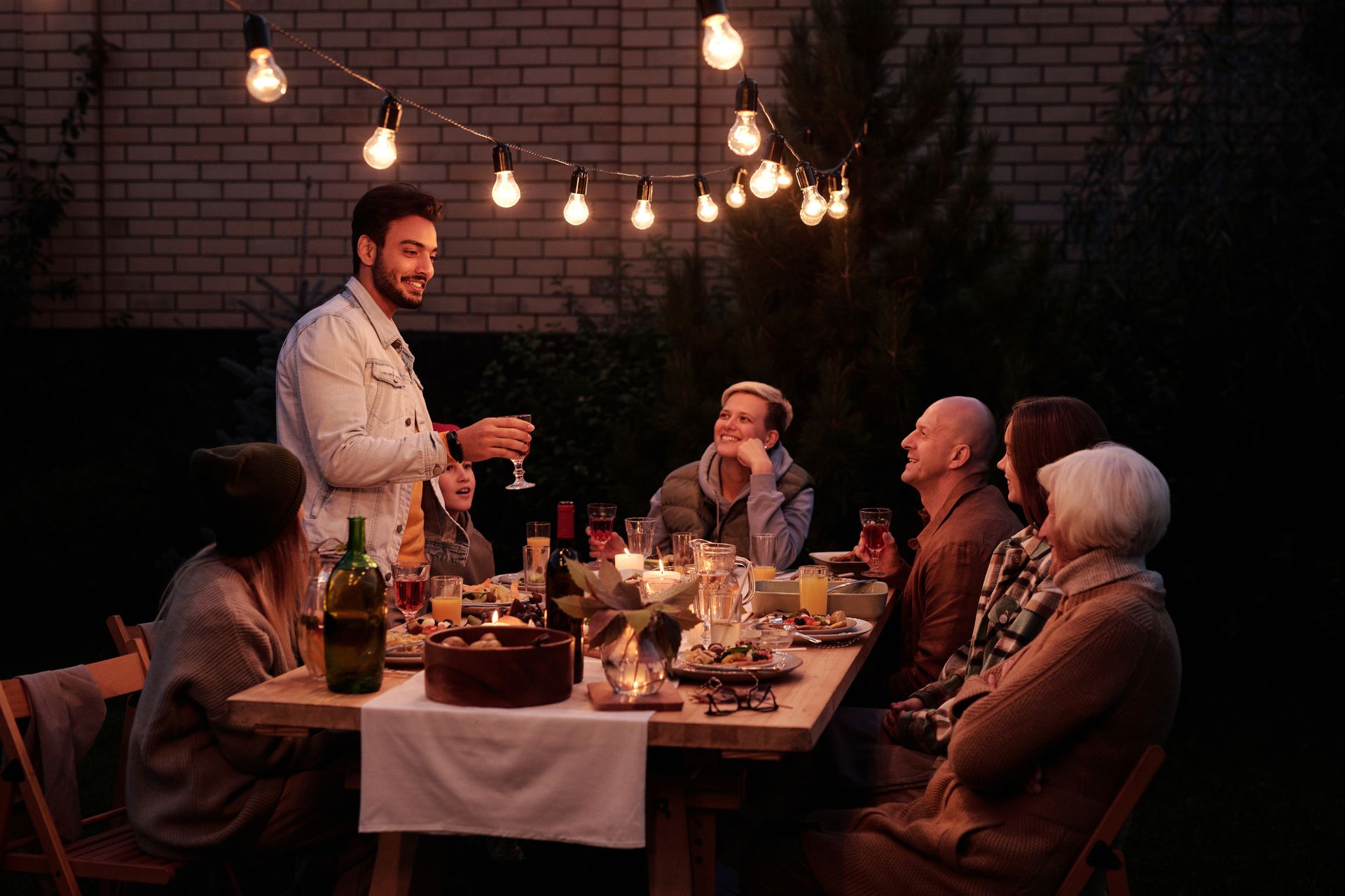 Neighbours share a garden dinner under string lights at dusk
